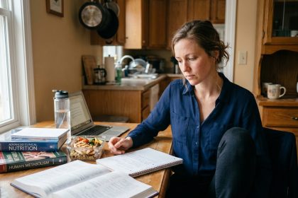 Femme adulte qui étudie le BTS diététique à la maison après sa journée de travail, entourée de livres de nutrition et de cours imprimés sur une table de cuisine